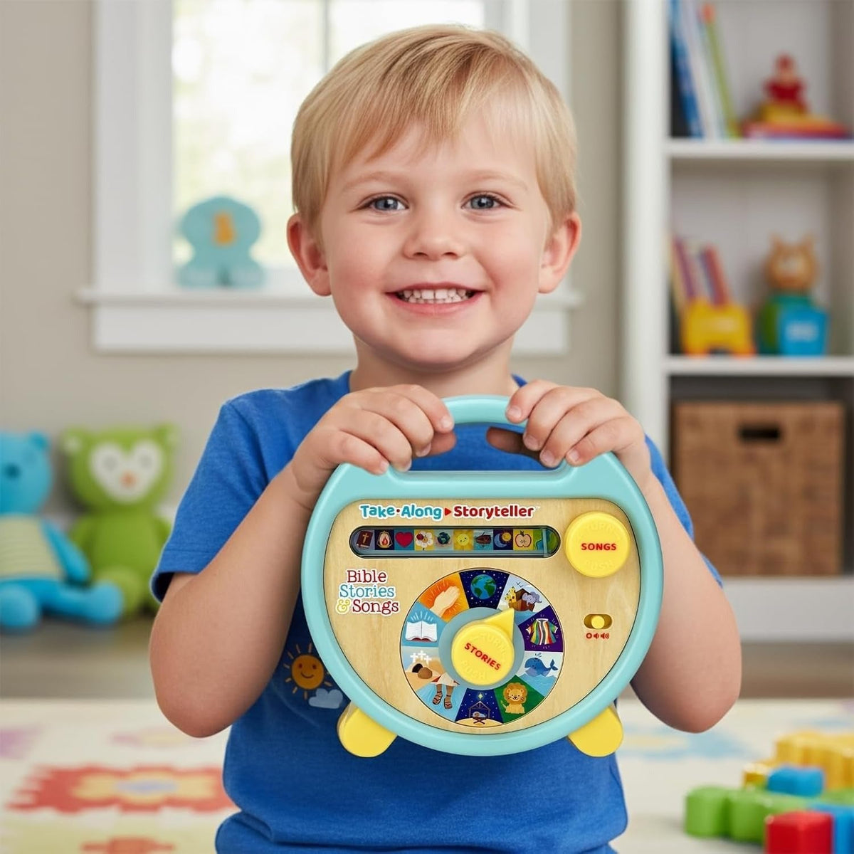 A smiling young boy in a blue shirt holds a colorful toy sing-along storytime player in a bright children's room. The round toy has a light blue handle and frame, a beige center panel, and yellow buttons and feet, with a multicolor dial showing different story elements.
